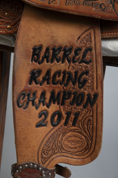 Close-up of a leather saddle with ornate tooling and engraved text reading "BARREL RACING CHAMPION 2011." There is a decorative pattern and a circular stamp on the leather.