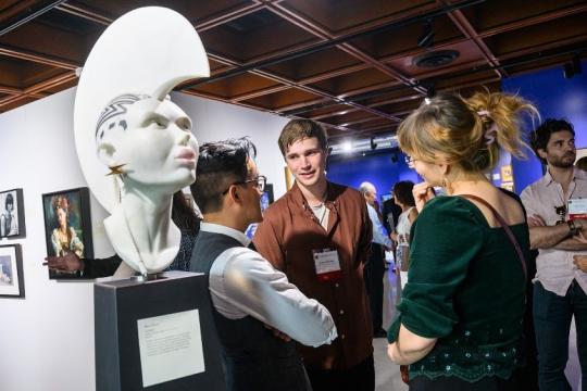 People converse in an art gallery near a sculpted bust with an elaborate hairstyle and facial markings. Paintings hang on the walls, and the setting is lively and modern.