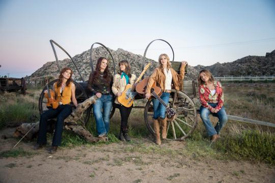 Five women pose outdoors in front of a rustic wooden wagon, with mountains in the background. One woman holds an electric guitar, and all are dressed in casual, country-style clothing.
