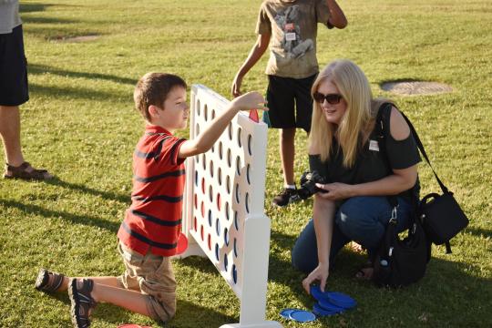A young boy kneels on the grass, playing a large outdoor Connect Four game with a smiling woman who is crouched nearby, holding blue game pieces. Another child stands in the background.