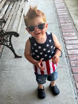 A young child with spiked hair wears star-spangled sunglasses, a USA flag tank top, denim shorts, and black shoes, smiling and standing on a sidewalk next to a wooden bench.