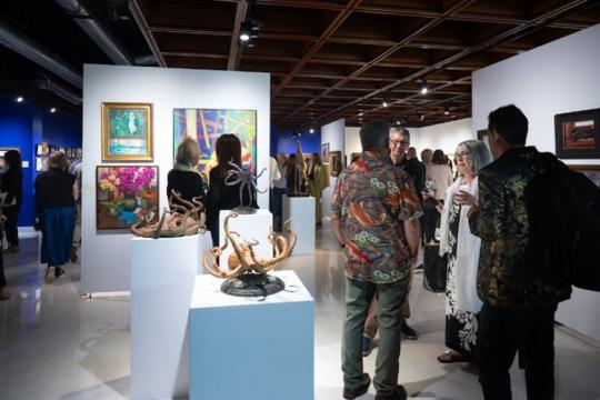 People gather and converse in an art gallery filled with paintings and sculptures, including detailed octopus sculptures exhibited on white pedestals. The space is brightly lit with a modern ceiling and white display walls.
