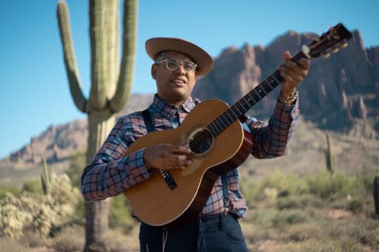 A man wearing glasses, a beret, and a plaid shirt plays an acoustic guitar in a desert landscape with cacti and mountains in the background.