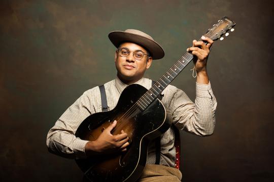 A man wearing glasses, a hat, and suspenders plays an acoustic guitar while looking at the camera, set against a brown and green mottled background.