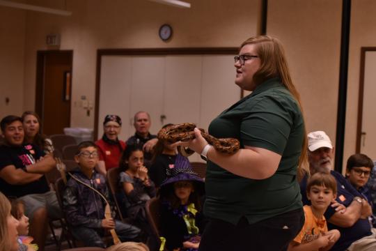 A woman stands in front of a seated audience holding a large snake. Children and adults watch attentively, some in costumes, in a room with beige walls and fluorescent lighting.