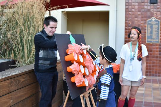 A man holds a board with orange cups shaped like a pumpkin while a child in a costume hits it with a stick. A woman in a costume stands nearby, watching. The scene appears to be part of a festive outdoor event.