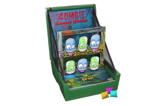 A tabletop carnival game called "Zombie Knock Down" features illustrated zombie faces on upright panels and three small colored beanbags in front for players to throw.
