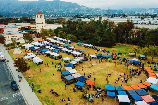 Aerial view of a community festival with colorful tents and booths set up on a grassy field, people walking around, trees nearby, and mountains in the background under an overcast sky.