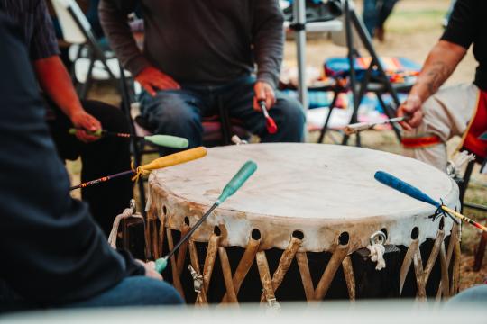 Four people seated around a large traditional drum, each holding a colorful drumstick and about to strike the drum’s surface, with blurred background items and chairs visible.