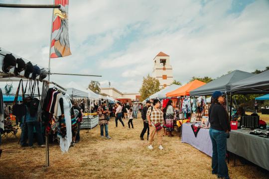 People browse vendor booths selling crafts, clothing, and jewelry at an outdoor market under tents. A tower building with a red roof is in the background, and the sky is partly cloudy.