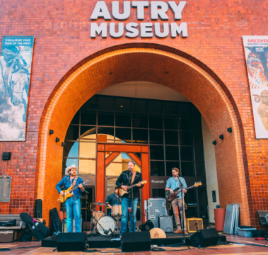 A band performs live music on an outdoor stage beneath the large arched entrance of the Autry Museum, with instruments, speakers, and museum signage visible in the background.