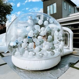 A large transparent inflatable dome filled with white and silver balloons sits outdoors on a patio, with a modern house and blue sky in the background.