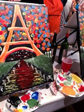 A colorful painting of the Eiffel Tower stands on an easel, surrounded by paint palettes, brushes, and bottles. Bright dabs of red, yellow, and blue fill the sky in the artwork, and people are painting in the background.
