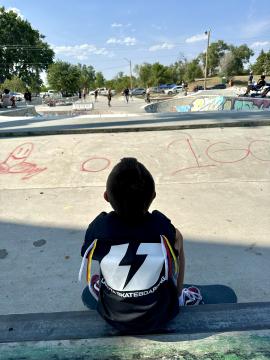 A child with short hair sits on a skateboard at a skatepark, wearing a black shirt with a large white lightning bolt on the back. People are skating in the background under a partly cloudy sky.