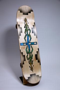 A skateboard deck with a geometric black border and a central design featuring green and blue shapes resembling stained glass, displayed upright on a stand against a plain background.