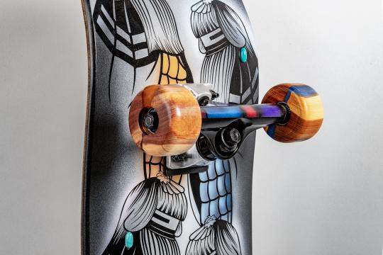 Close-up of a skateboard showing colorful wooden wheels, a metallic truck, and deck artwork featuring stylized black-and-white illustrations of figures with long hair and turquoise accents.