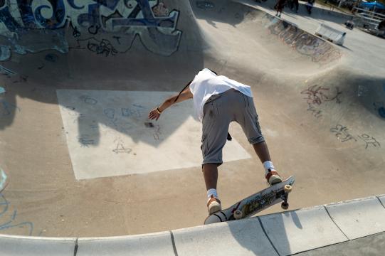 A skateboarder in a white shirt and gray shorts prepares to drop into a concrete skatepark bowl covered in graffiti and drawings, captured from behind under bright daylight.
