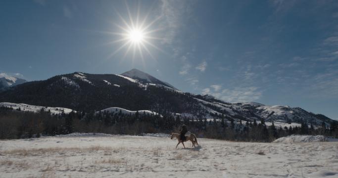 A person rides a horse across a snowy field with snowy mountains and pine trees in the background, while the sun shines brightly in a clear blue sky.