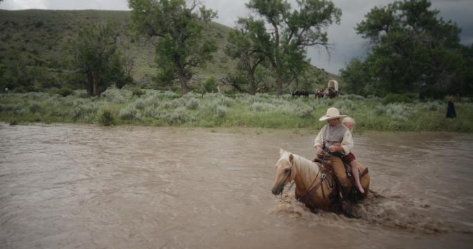 A person wearing a cowboy hat rides a light brown horse through a muddy river, carrying a child in front. Green hills and trees are in the background under a cloudy sky.