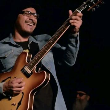 A smiling person wearing glasses, a hat, and a denim jacket plays an electric guitar on stage. Another person with a beard and sunglasses is visible in the dark background.