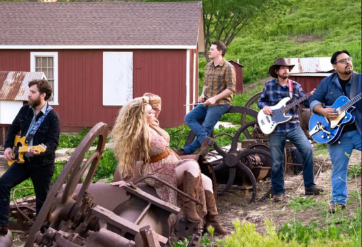 Five musicians with guitars and a drum sit and stand among old farm equipment in a grassy outdoor setting, with a red barn and green trees in the background. One woman is seated in front, while four men play instruments around her.