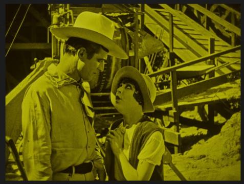 A man in a cowboy hat and a woman in vintage clothing stand close together, looking at each other with serious expressions, against a backdrop of industrial machinery and stairs tinted yellow.