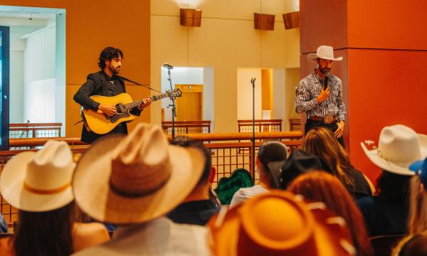 A musician plays guitar and sings on stage while a man in a cowboy hat and large belt buckle gestures to an audience, many of whom are also wearing cowboy hats, in an indoor venue with warm lighting.