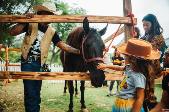 A young girl in a cowboy hat pets a dark horse through a wooden fence, surrounded by adults and children at an outdoor event. Trees and a white picket fence are visible in the background.