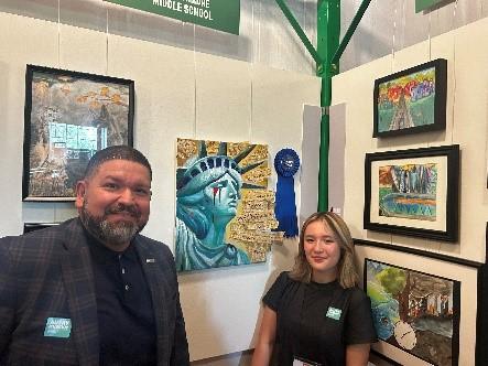A man and a young woman stand in front of an art display featuring paintings, including one of the Statue of Liberty with a blue ribbon award. Both are smiling and wearing name badges.