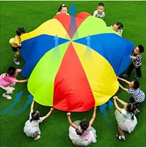 Children stand in a circle on green grass, holding and lifting a large, colorful parachute with red, yellow, green, and blue sections. The parachute billows upward as they play together.