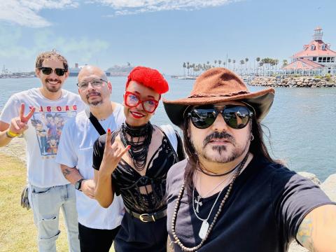 Four people pose for a selfie near a waterfront with palm trees and a red-roofed building in the background. They appear cheerful, wearing casual and stylish outfits, with sunny weather overhead.