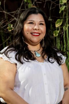 A woman with long dark hair and red lipstick smiles outdoors. She wears a white blouse with lace trim, turquoise pendant necklace, and earrings. Green plants and vines are in the background.