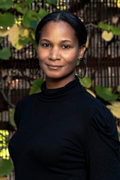 A woman wearing a black long-sleeve top and hoop earrings stands outdoors in front of a background with green leaves and a brown fence, looking at the camera with a gentle smile.