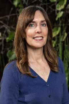 A woman with long brown hair and blue eyes, wearing a blue buttoned sweater and a necklace, smiles in front of a blurred outdoor background with green leaves and branches.