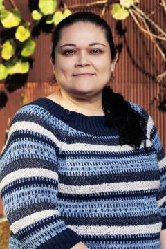 Woman with dark hair pulled back, wearing a blue and white striped sweater, stands outdoors in front of a brown fence with green leaves and branches in the background, smiling gently at the camera.