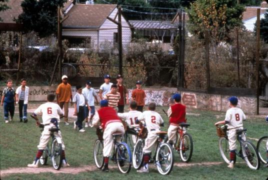 A group of boys on bikes wearing baseball uniforms face another group of kids standing on a grassy baseball field, with houses and trees in the background.