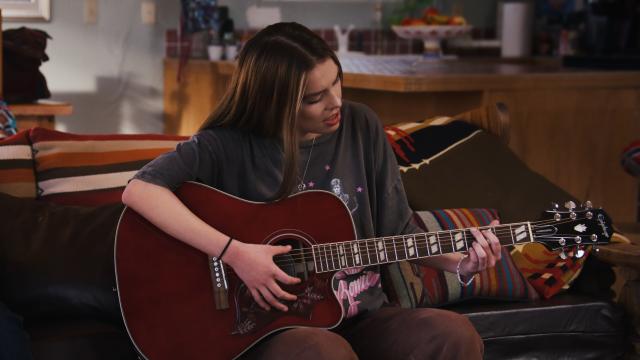 A young woman sits on a couch, playing an acoustic guitar. She is casually dressed in a gray t-shirt and brown pants, focused on strumming the instrument in a cozy, warmly lit living room.