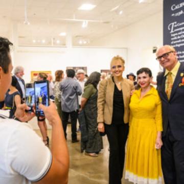 A person takes a photo with a smartphone of three people posing together at an art gallery event; many other attendees are mingling in the brightly lit gallery space.