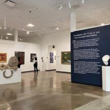 Art gallery interior with sculptures on pedestals and paintings on white walls. A large dark blue wall displays exhibition text. Two people are viewing the artworks in the background. The floor is polished concrete.