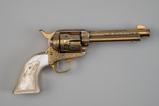 A decorative revolver with intricate gold engravings and a white handle featuring an eagle design with a blue eye, displayed on a plain gray background.