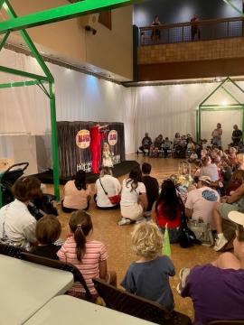 A performer in a long red dress stands on stage in front of a seated audience of children and adults inside a brightly lit indoor space with white decorations and green structures.