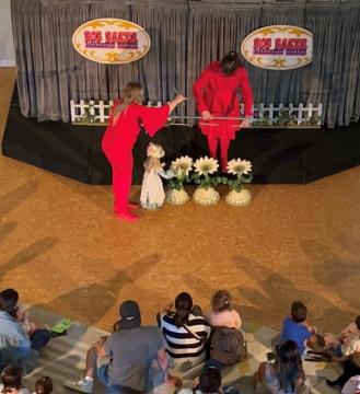 Two puppeteers in red outfits perform with a marionette dressed in white on a stage decorated with large artificial sunflowers, as a seated audience of children and adults watches.