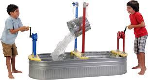 Two boys play with a water activity table, each operating a hand pump that tips a metal bucket to pour water into a large metal trough.