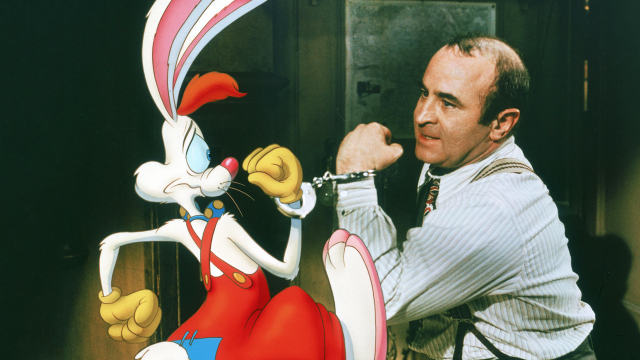 A man in a striped shirt is handcuffed to a cartoon rabbit with red overalls and long ears. The rabbit looks anxious while the man seems frustrated. They are indoors.