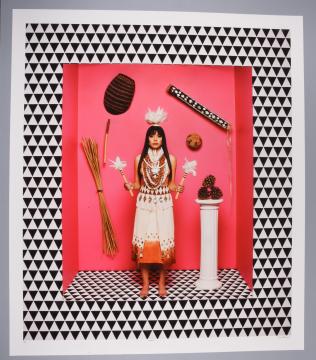 A woman in traditional dress stands holding feathers, surrounded by natural objects on a pink background, framed by a black and white triangle pattern.