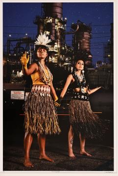 Two women in traditional Polynesian attire stand in front of a lit industrial refinery at night, wearing grass skirts, jewelry, and headdresses, with their hands raised and painted yellow.