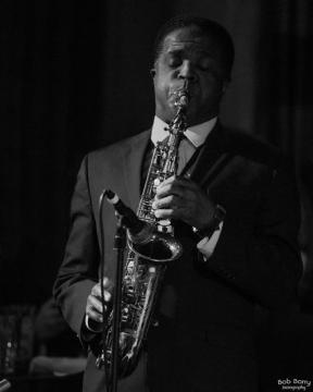 A man in a suit plays a saxophone passionately on stage. The image is black and white, with dramatic lighting highlighting his focused expression. A microphone is positioned in front of him.