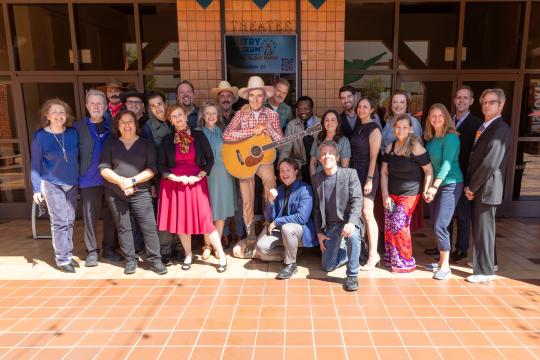 A group of about 22 adults pose together outside a building; one person in the center holds a guitar and wears a cowboy hat. Most people are smiling and standing on tile flooring in front of glass doors.