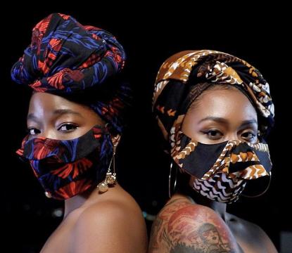two women standing back to back looking at the camera both wearing headdress with matching mask, black background