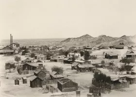 black and white photograph of a small town in the desert, with dirt hills in the background of the buildings